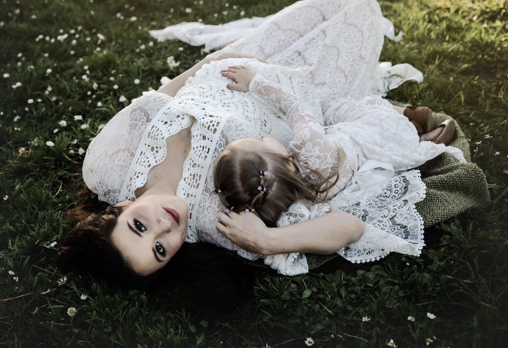 Woman and child in white lace outfits, lying in a field of green grass and small white flowers.