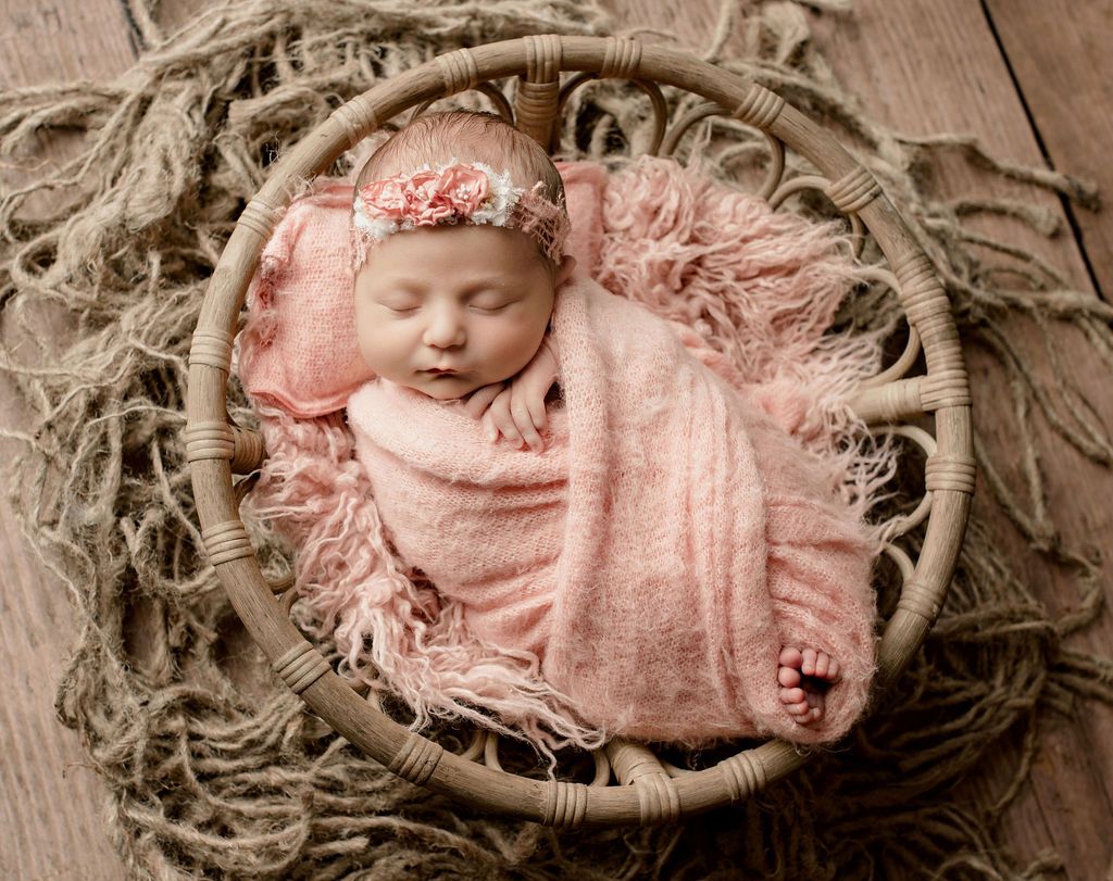 Newborn baby swaddled in pink, asleep in a woven basket surrounded by rope.