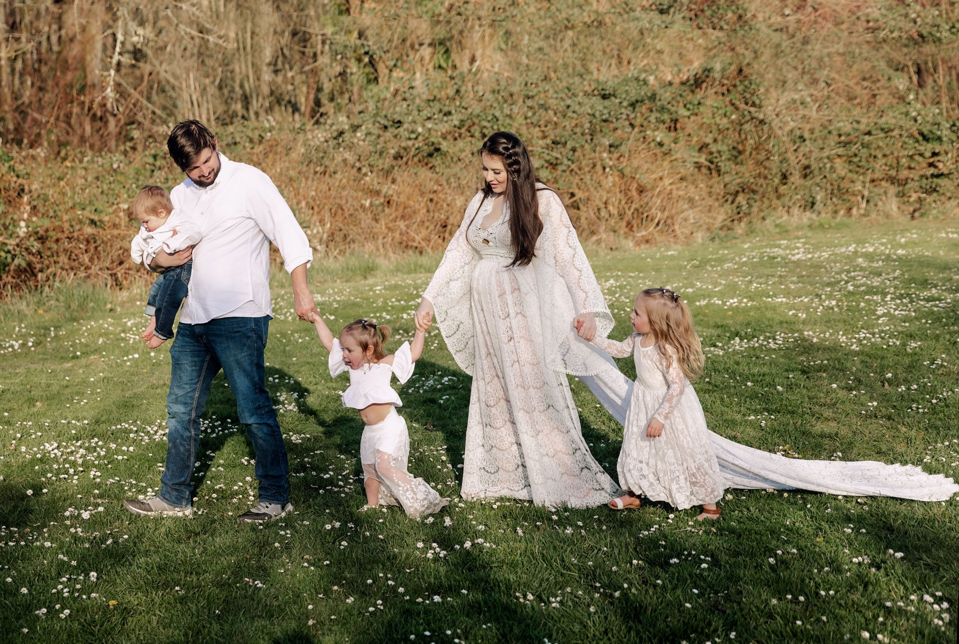 Family of five walking on grass, two young girls holding parents' hands, boy held.