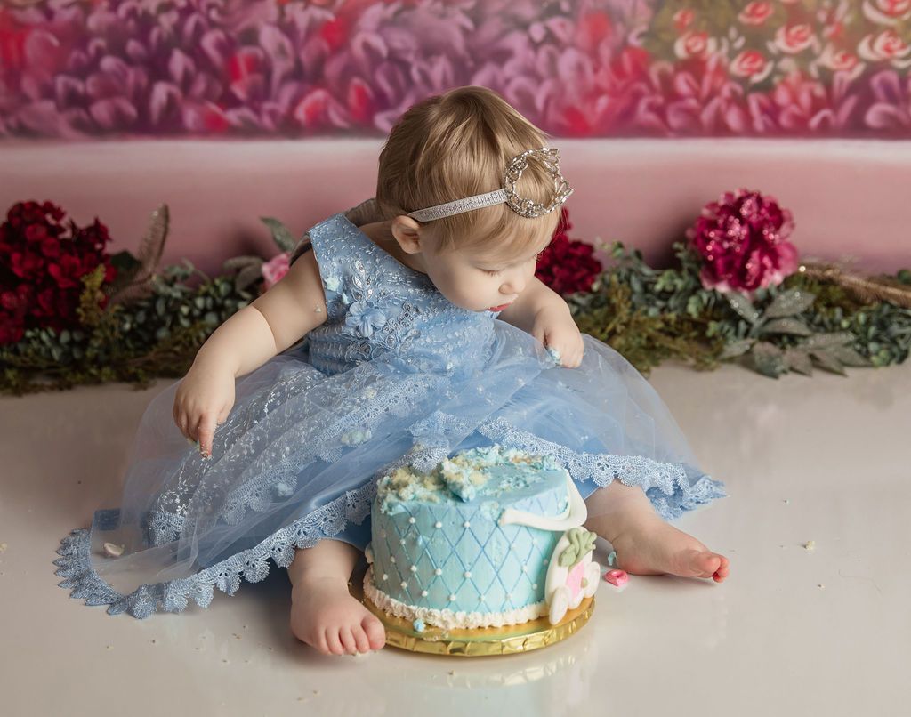 Baby in blue dress reaches for cake on a golden platter with floral backdrop.
