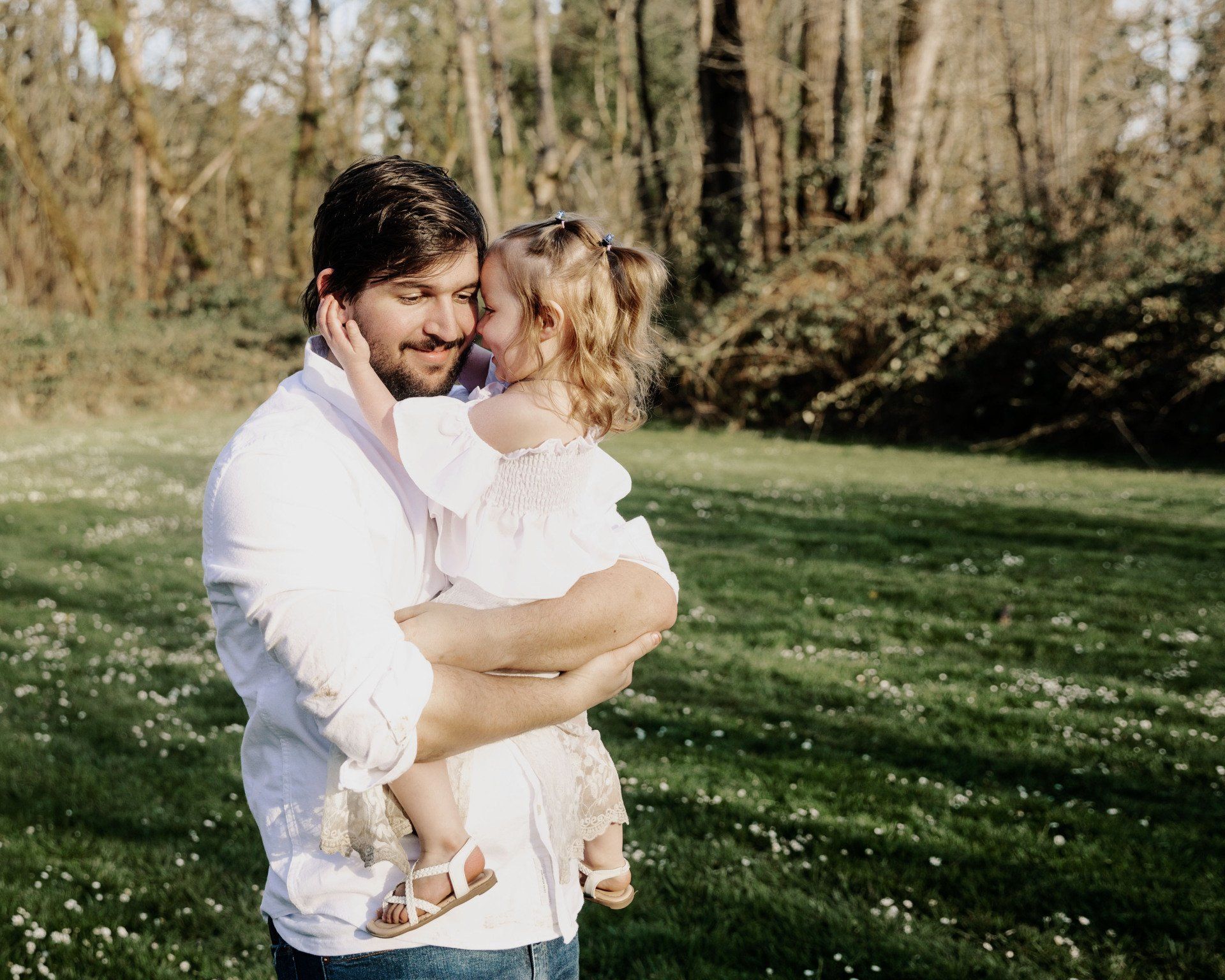 Man holds child, smiling and embracing in grassy field, trees in the background.
