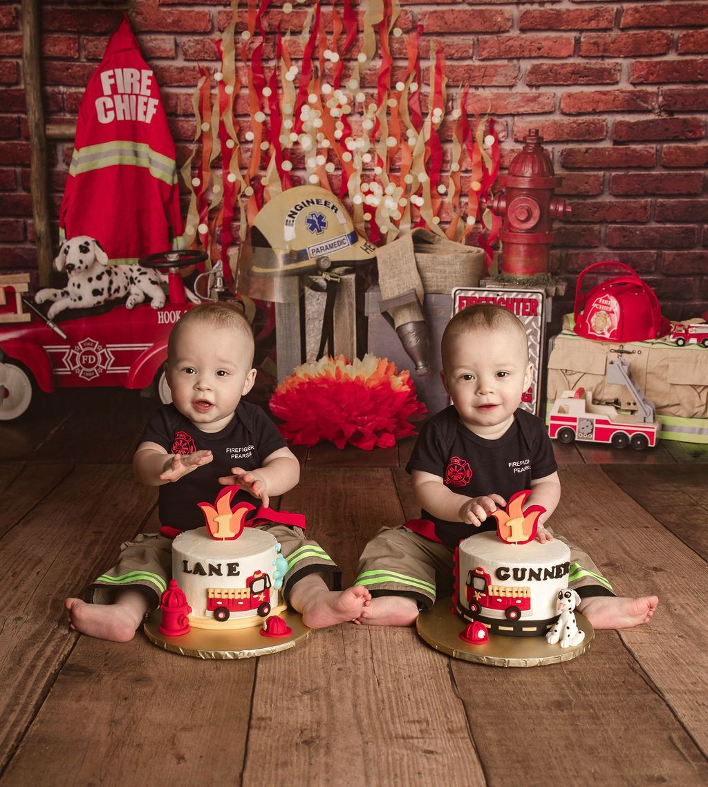 Twins in firefighter-themed outfits sitting in front of cakes. Fire-themed decorations and backdrop.