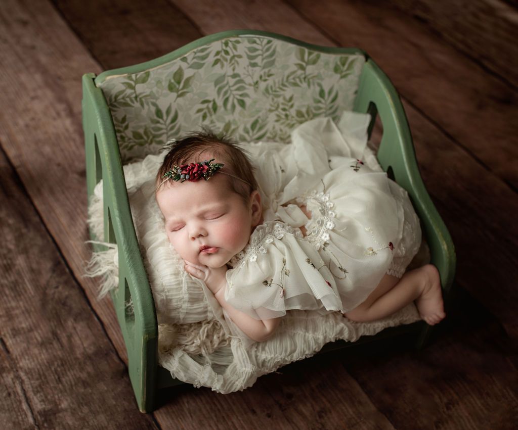 Newborn baby sleeping in a small green bed with floral print, on a cream blanket, wooden floor.