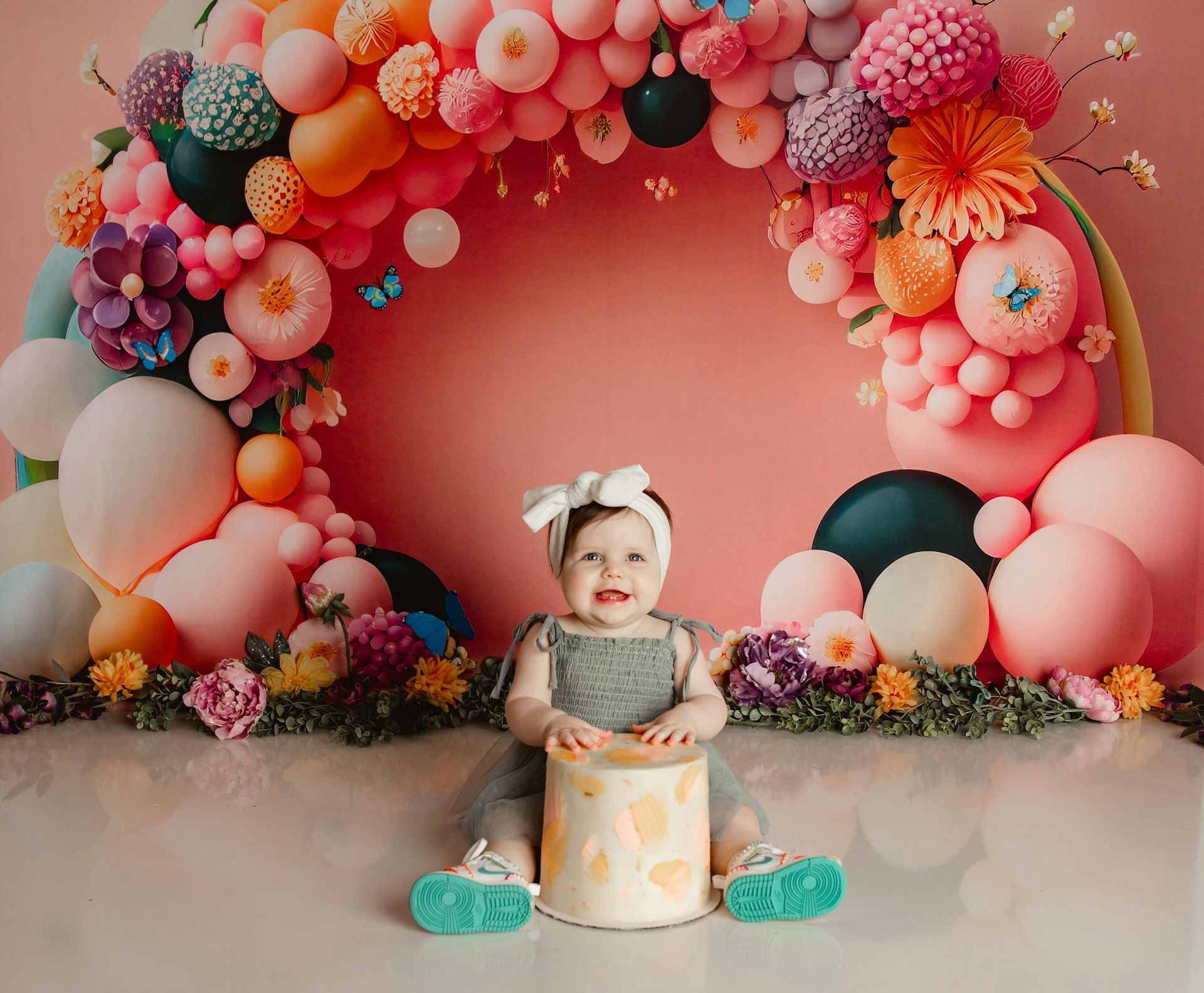 Smiling baby with cake, in front of a colorful balloon arch and floral decorations.