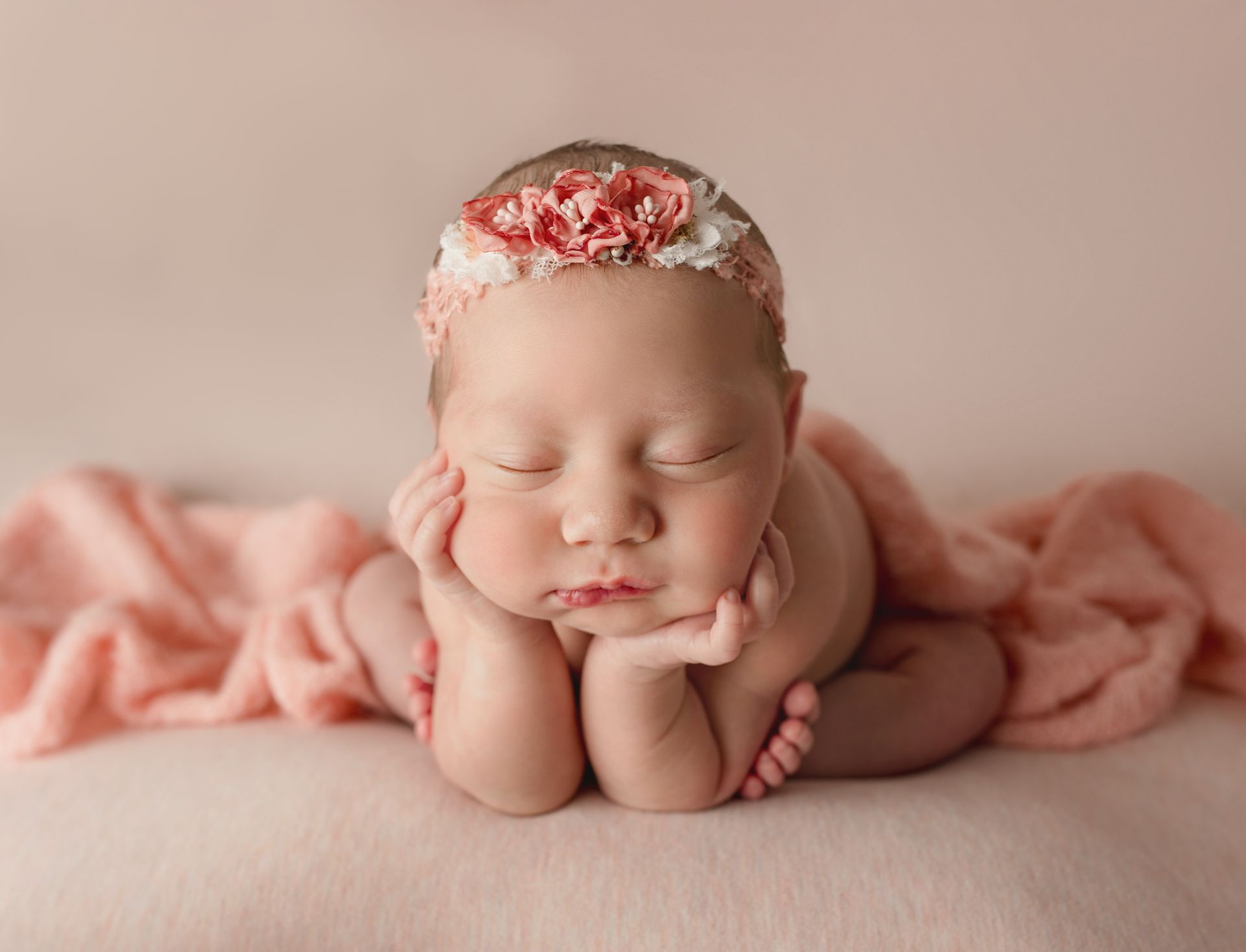 Newborn baby, eyes closed, resting on hands, wearing a floral headband, wrapped in pink blanket.