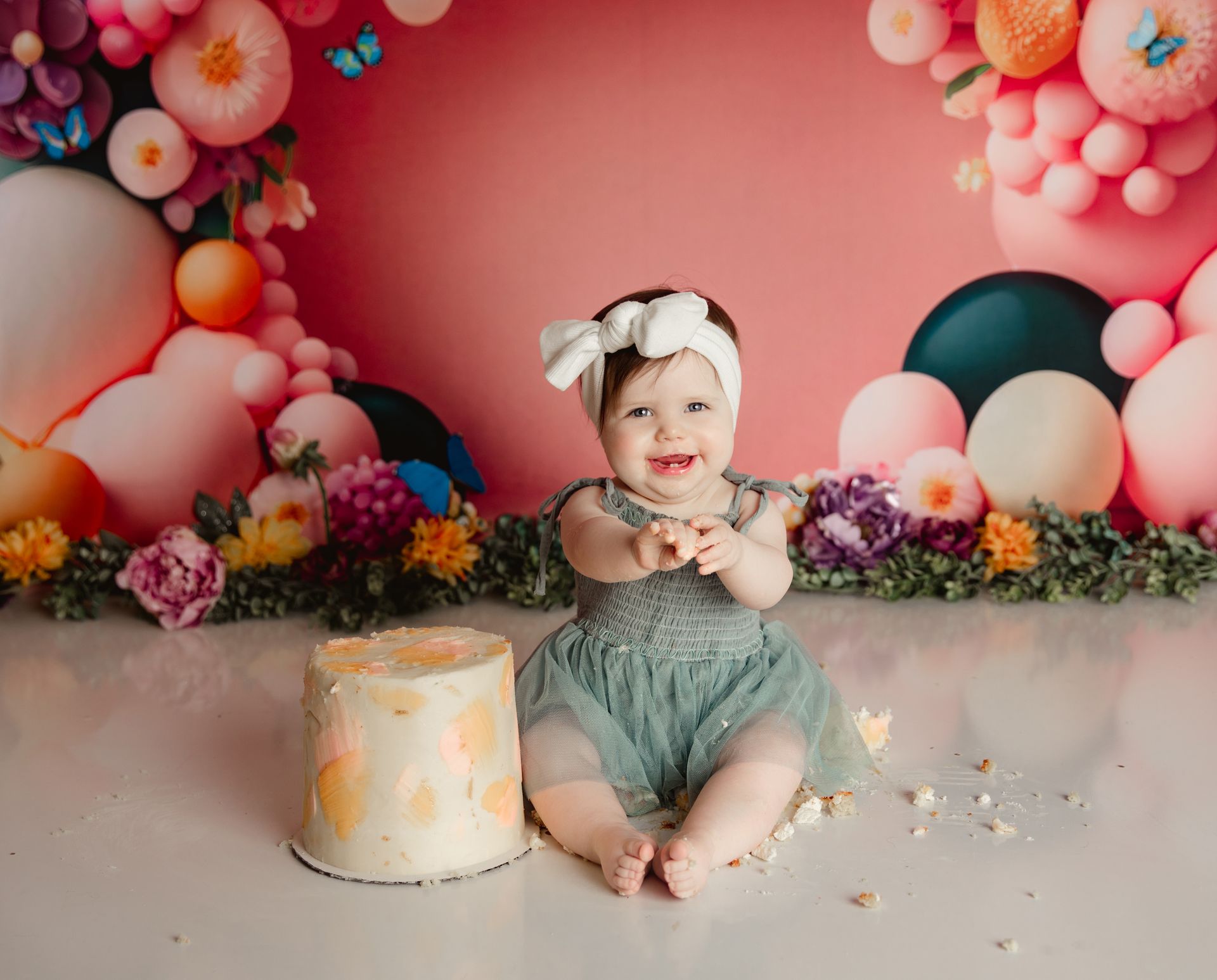 Smiling baby in a blue dress with a white bow, seated in front of a cake and a balloon arch.