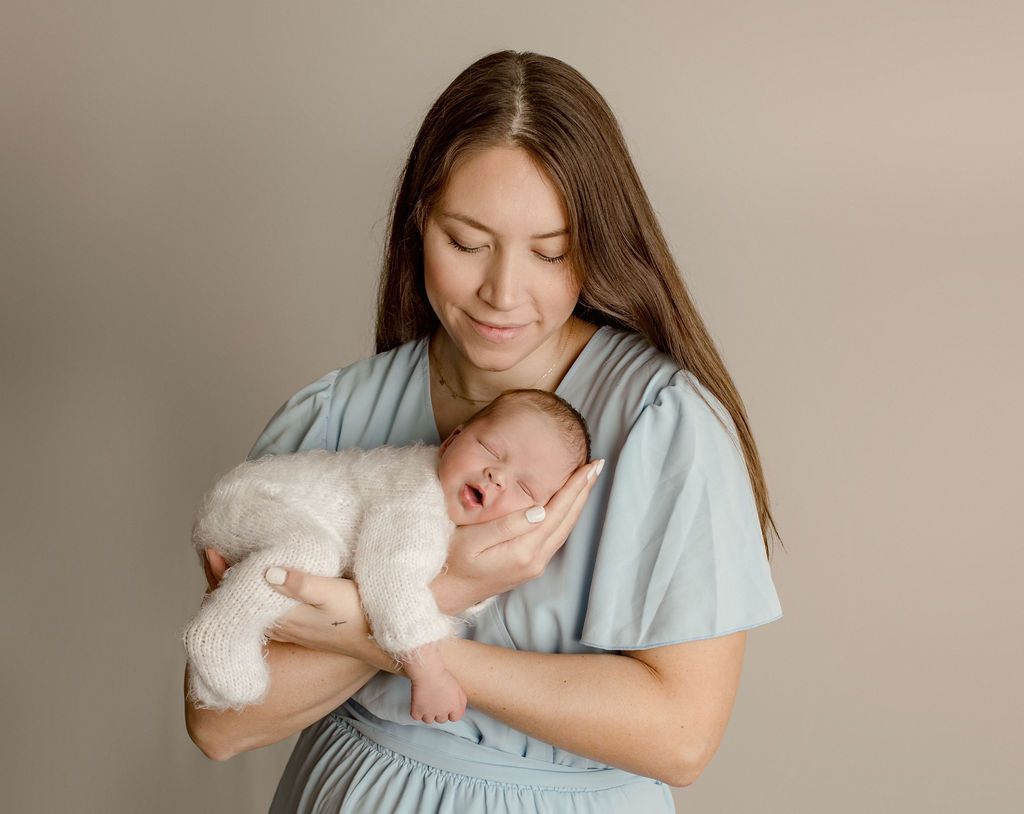 Woman in a blue dress cradles a sleeping newborn in a white outfit, against a neutral background.