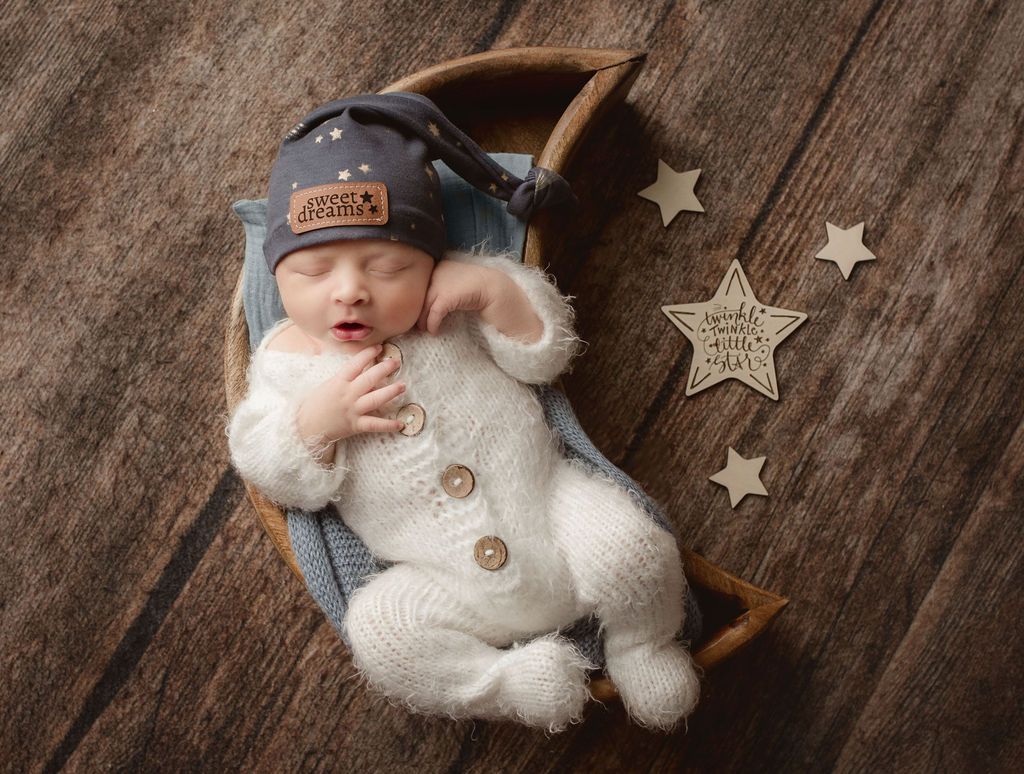 Newborn baby in a white knit outfit and nightcap, asleep in a wooden crescent moon.
