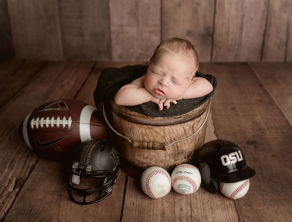 Sleeping baby in a wooden bucket surrounded by sports equipment: football, baseballs, and a helmet.