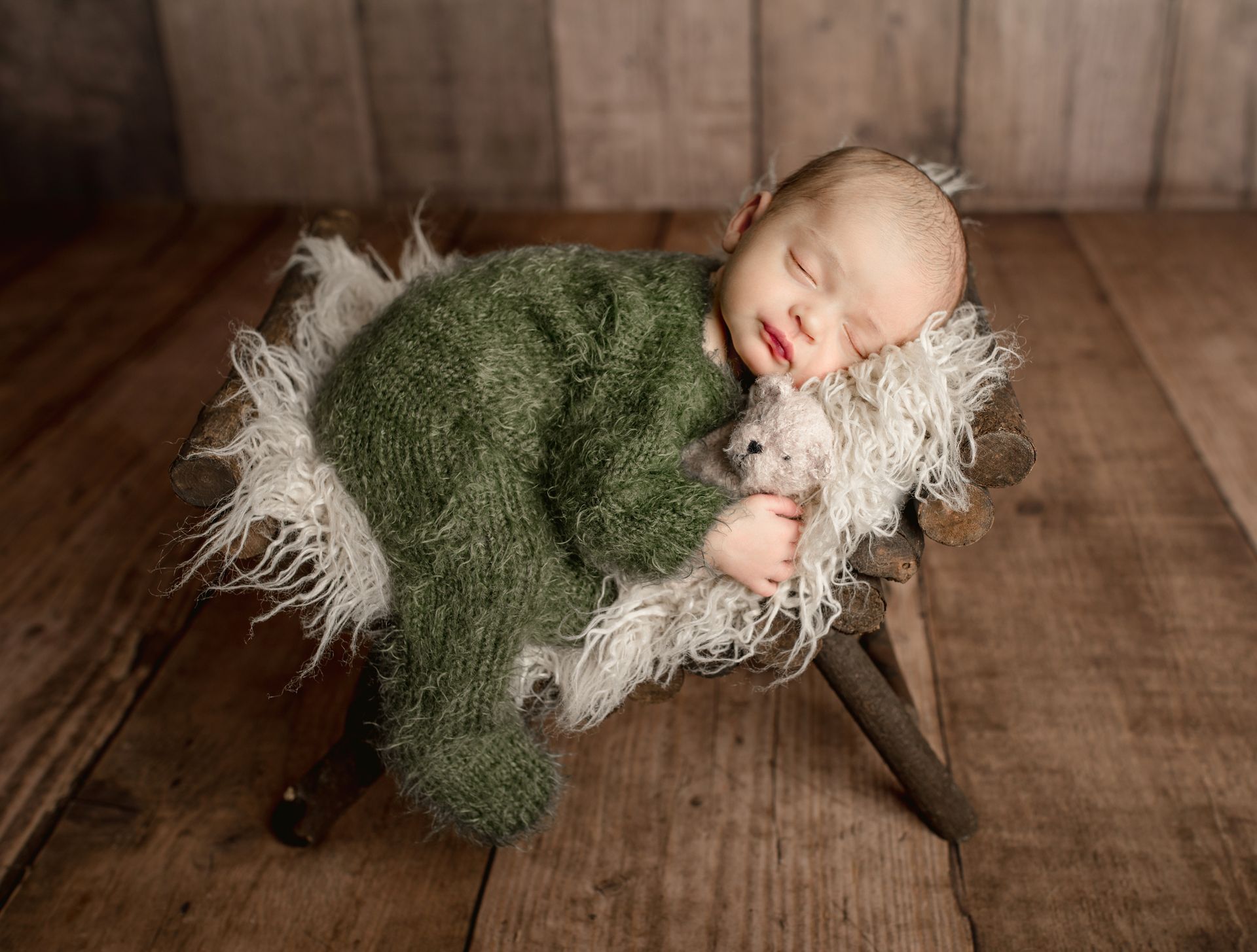 Sleeping baby in green outfit, curled in a wooden cradle with white fluff, on wooden planks.
