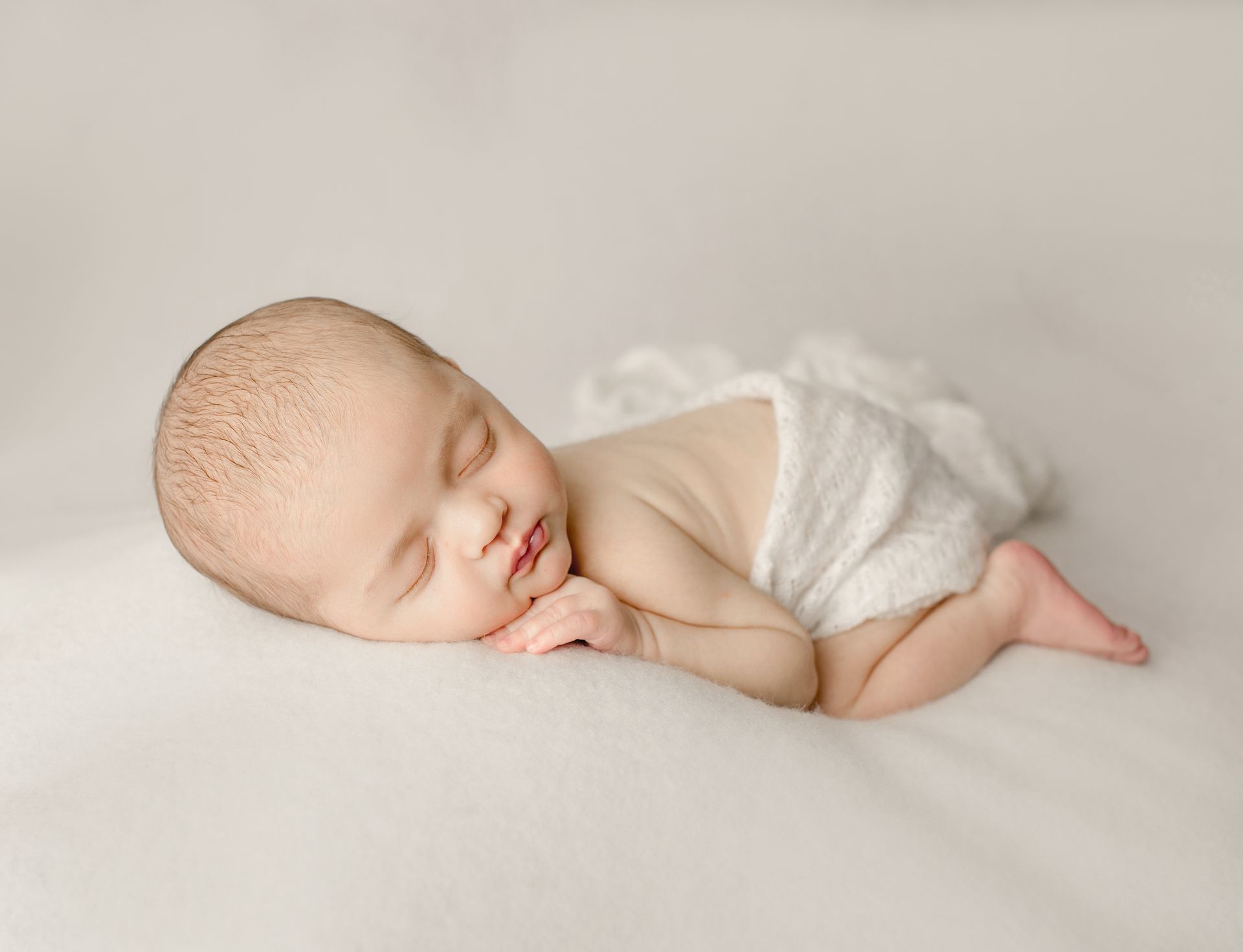Sleeping newborn baby on a white blanket, wearing a swaddle.