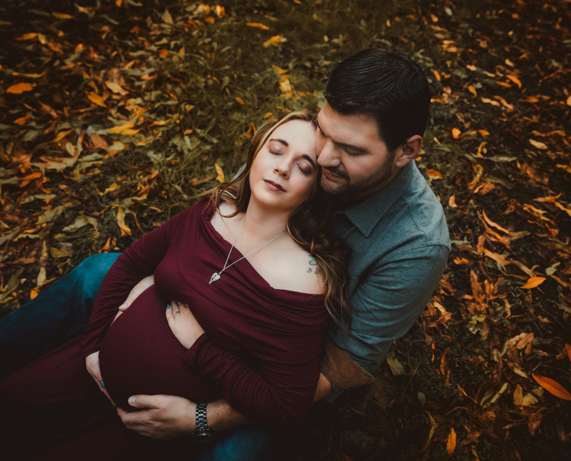 Pregnant person in burgundy dress rests head on partner's shoulder, they sit amidst autumn leaves.