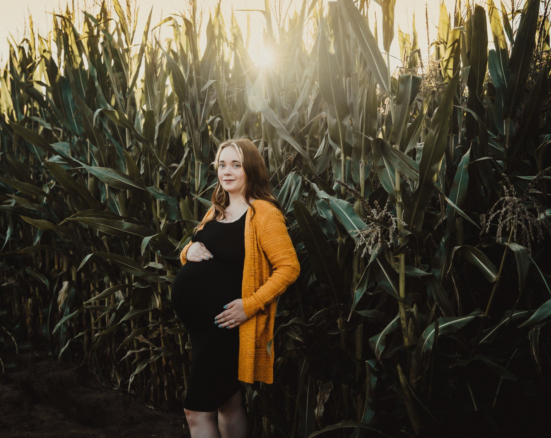Pregnant woman in black dress and yellow cardigan stands in front of corn stalks; sunlight shines.