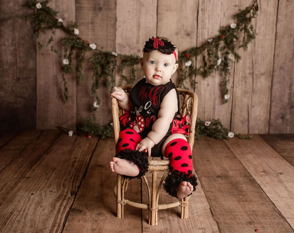 Baby in ladybug costume sits on wicker chair against wooden backdrop.