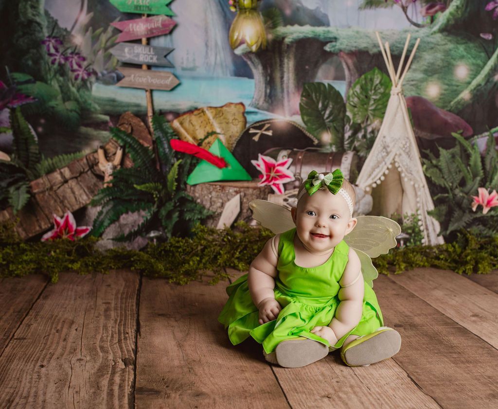 Baby dressed as a fairy with green dress and wings, smiling on wooden floor with fairytale backdrop.