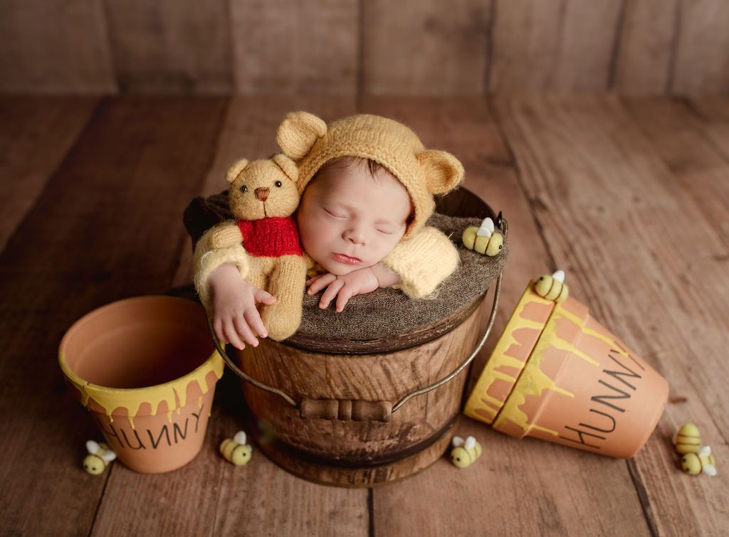 Newborn in Winnie-the-Pooh costume sleeps in a wooden bucket with Pooh bear toy, surrounded by honey pots and bees.