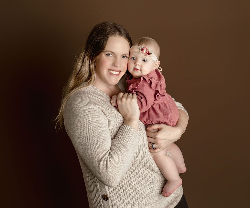 Woman in beige sweater holds a baby in a pink dress, both smiling, against a brown background.