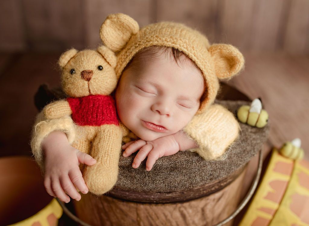 Newborn in Winnie the Pooh costume, sleeping in a wooden bucket with a stuffed bear.