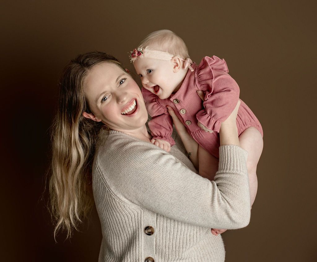 Woman holding smiling baby in front of a brown backdrop; both are looking up and smiling.