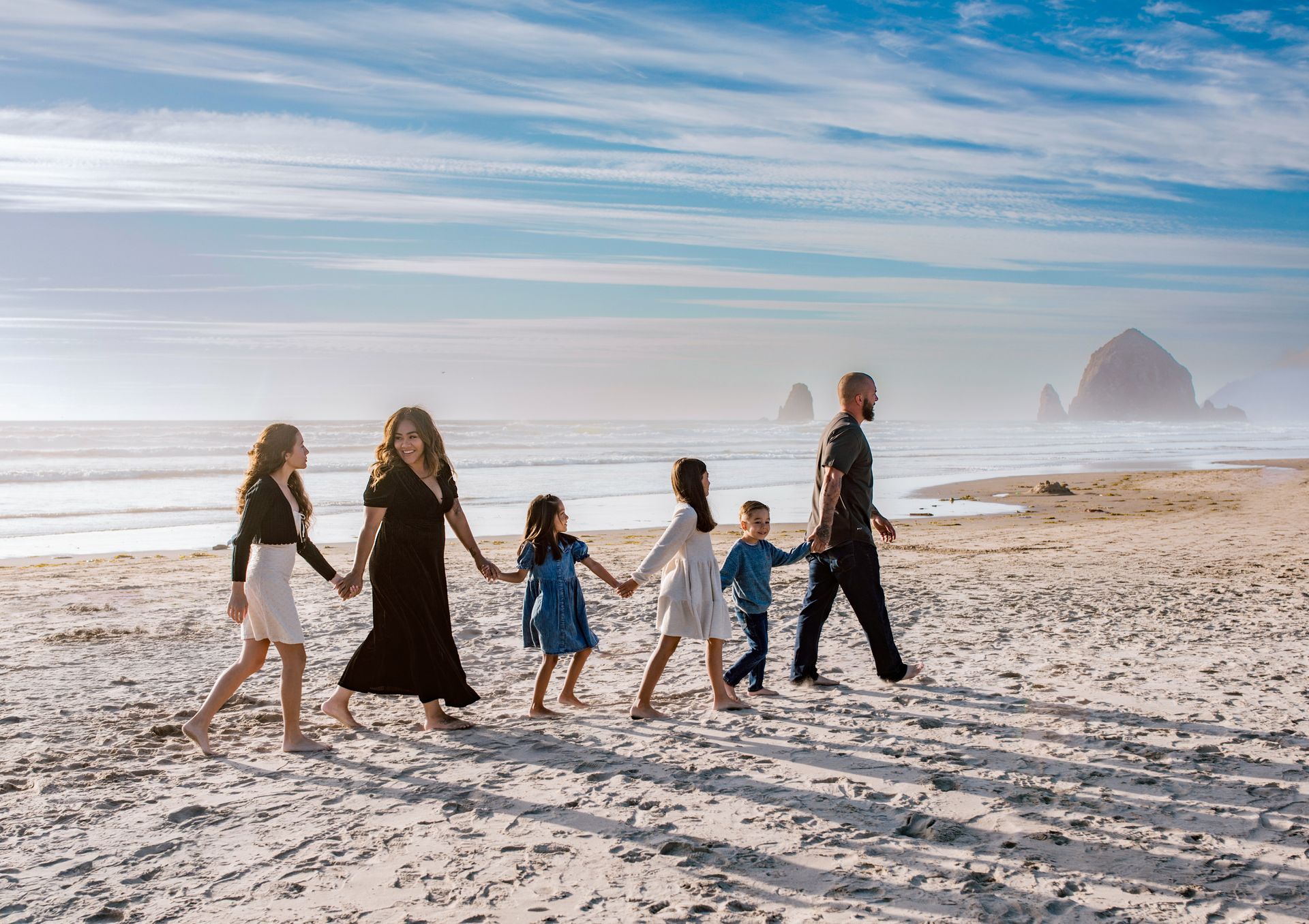 Family walking on a sandy beach, holding hands. Ocean and cloudy sky in the background.