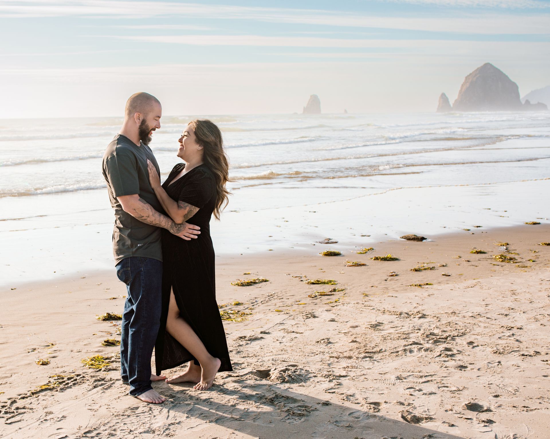 Couple embracing on a beach, smiling. The ocean and large rock formations are in the background.