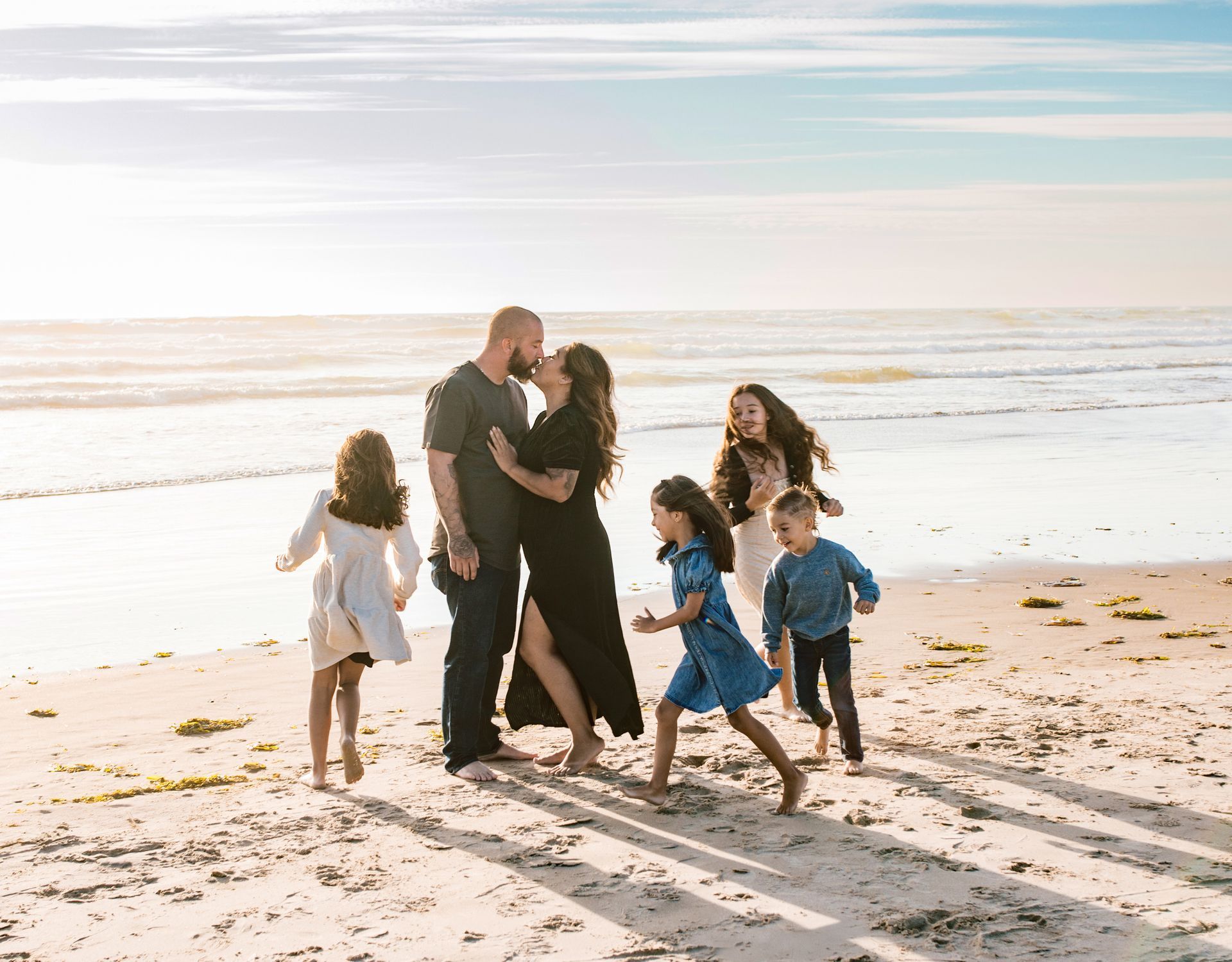 Family kissing on a beach. Children run nearby. Sunny day, ocean in the background.