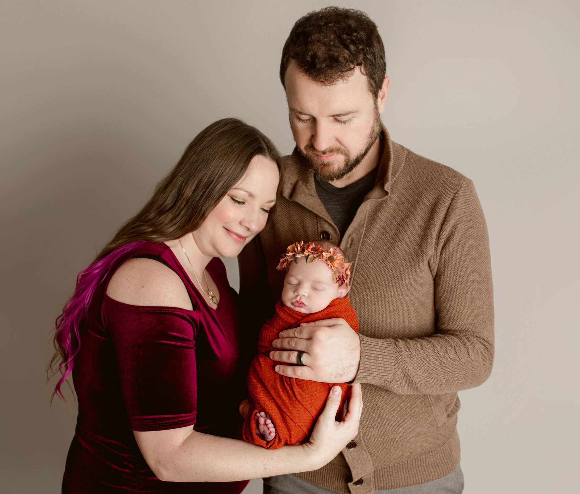 Couple holding a newborn baby wrapped in a red blanket; posing against a gray backdrop.