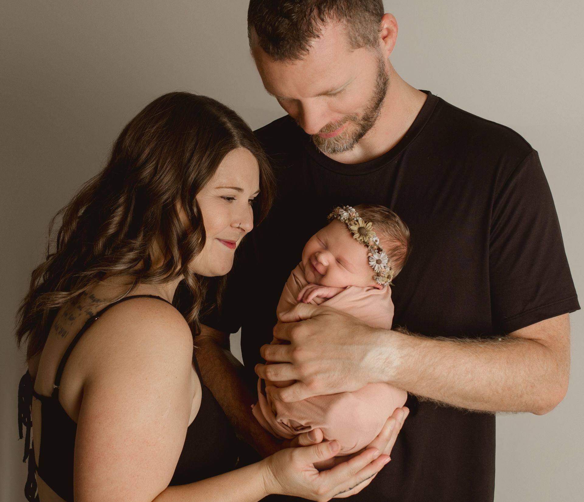 Parents holding newborn, baby smiling, neutral background.