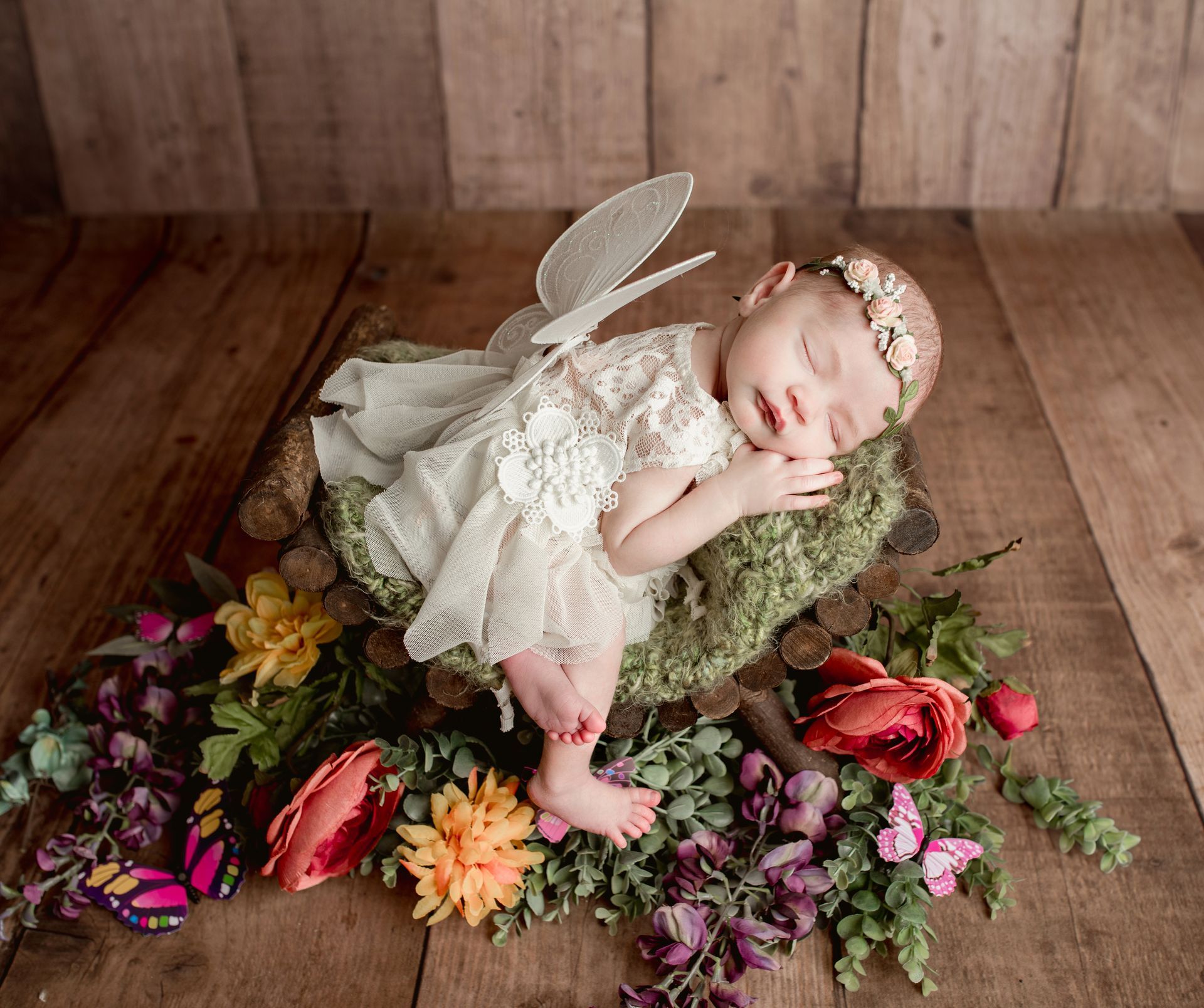 Newborn in fairy costume, sleeping in moss nest with flowers, wings, and wooden background.