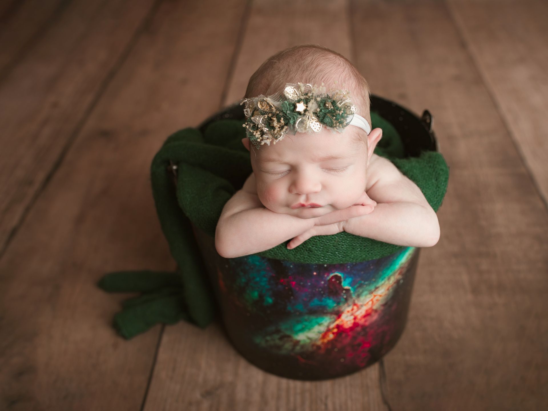 Newborn baby sleeping in a painted bowl, wrapped in green blanket, wearing floral headband, on wooden floor.