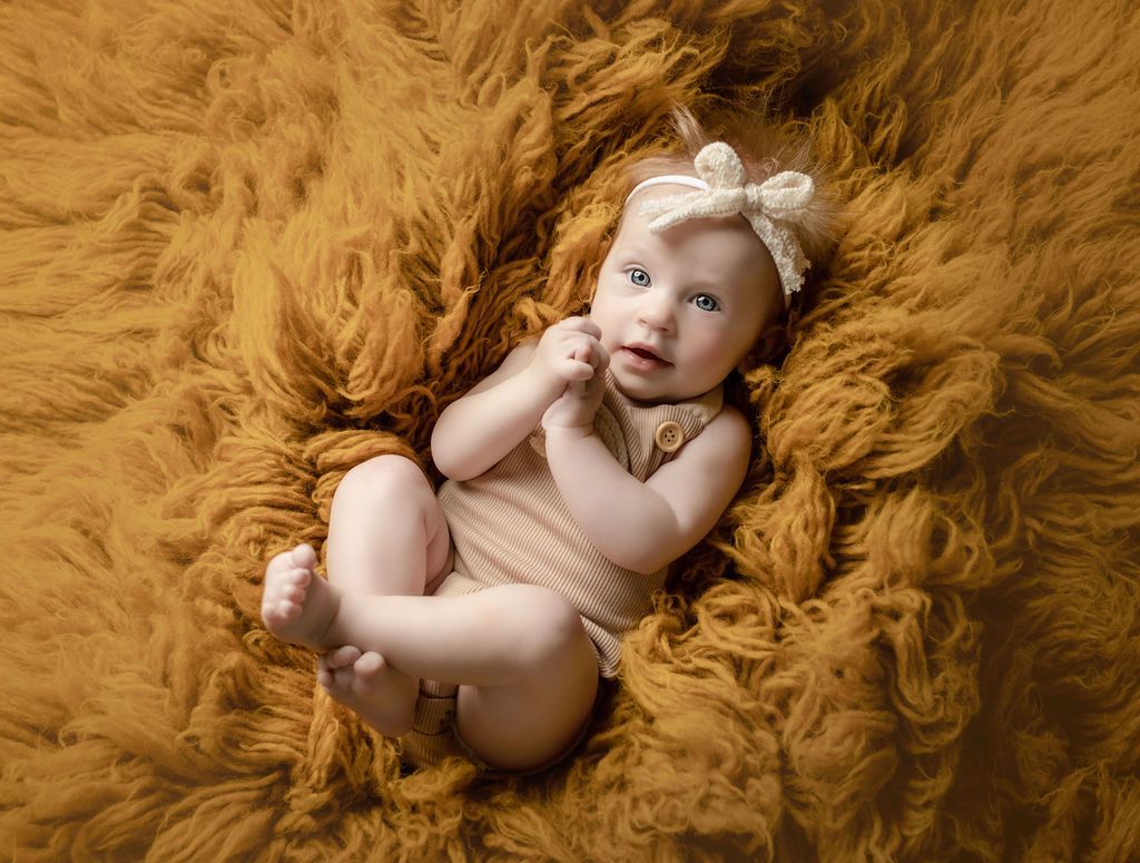 Baby in beige bodysuit and bow headband, resting on a fluffy, rust-colored blanket.