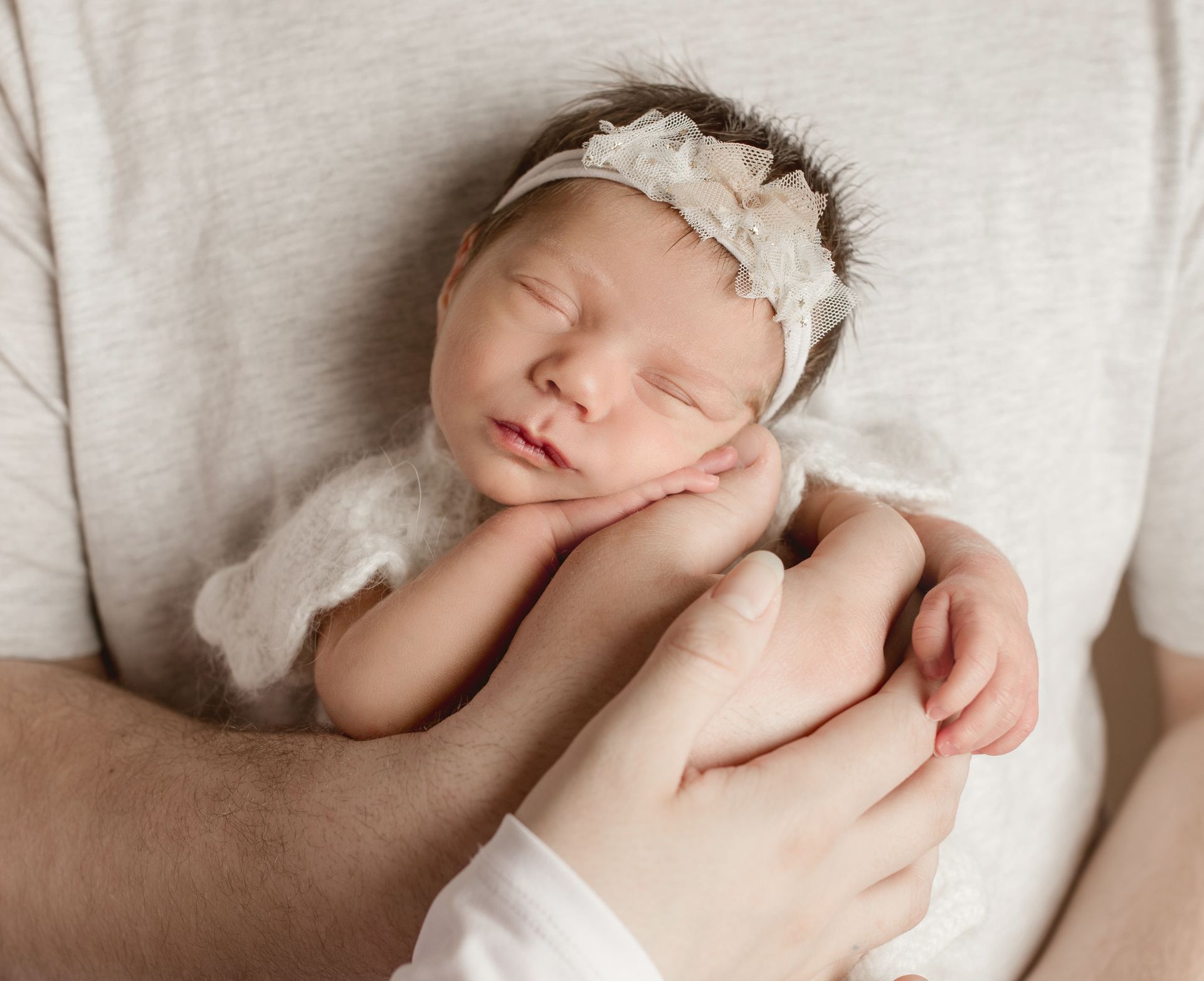 Newborn baby sleeping, held gently in someone's arms. Baby wears a lace headband and light-colored outfit.