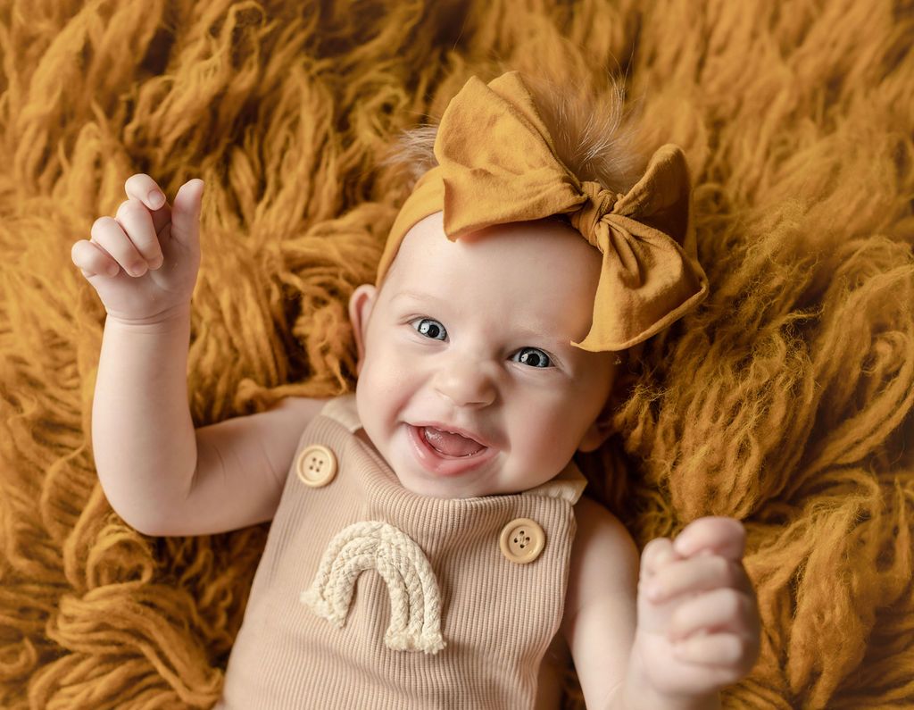 Smiling baby wearing mustard bow and jumpsuit, lying on fuzzy mustard blanket.