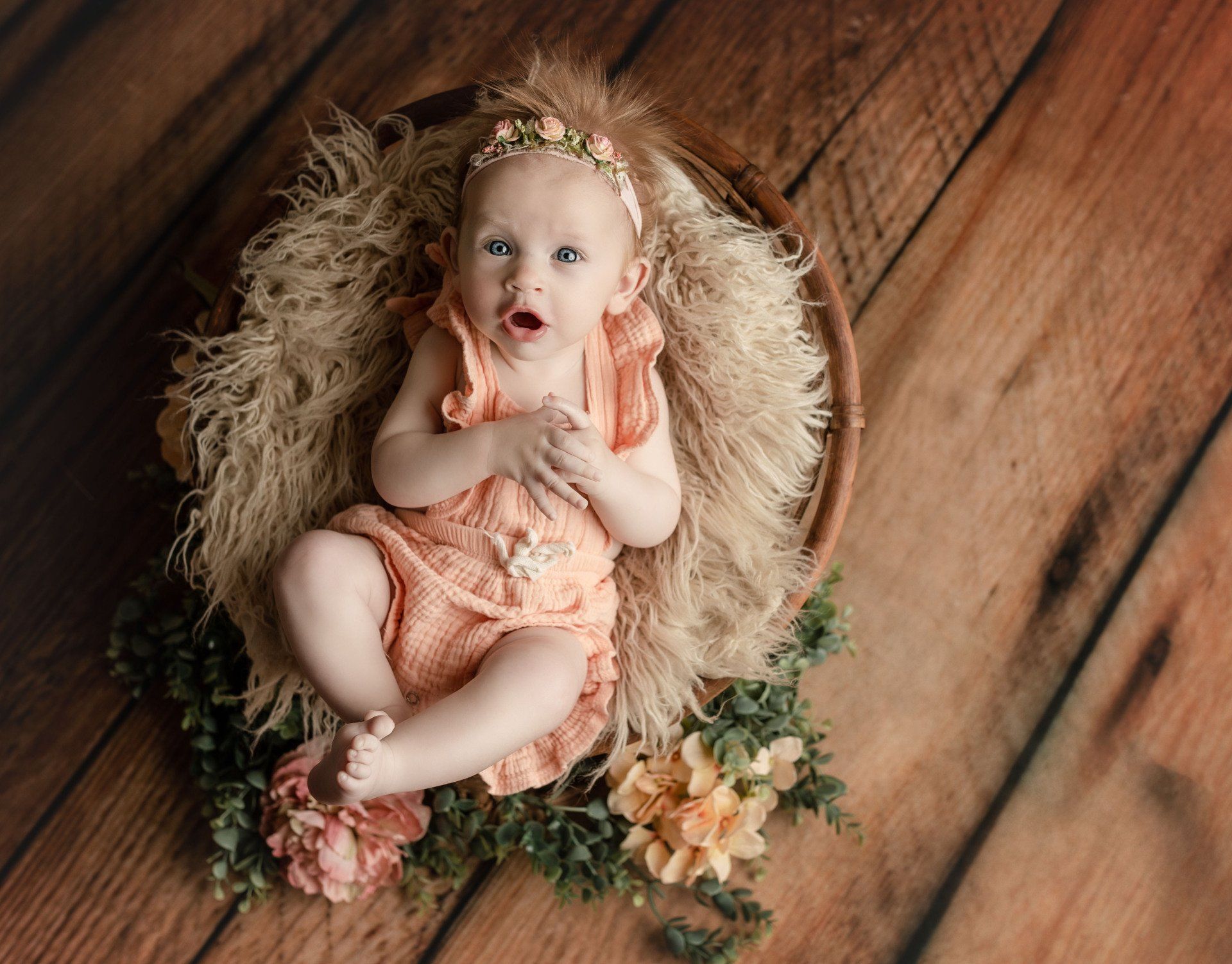 Baby in peach outfit lies in a nest of straw and flowers on a wooden floor.