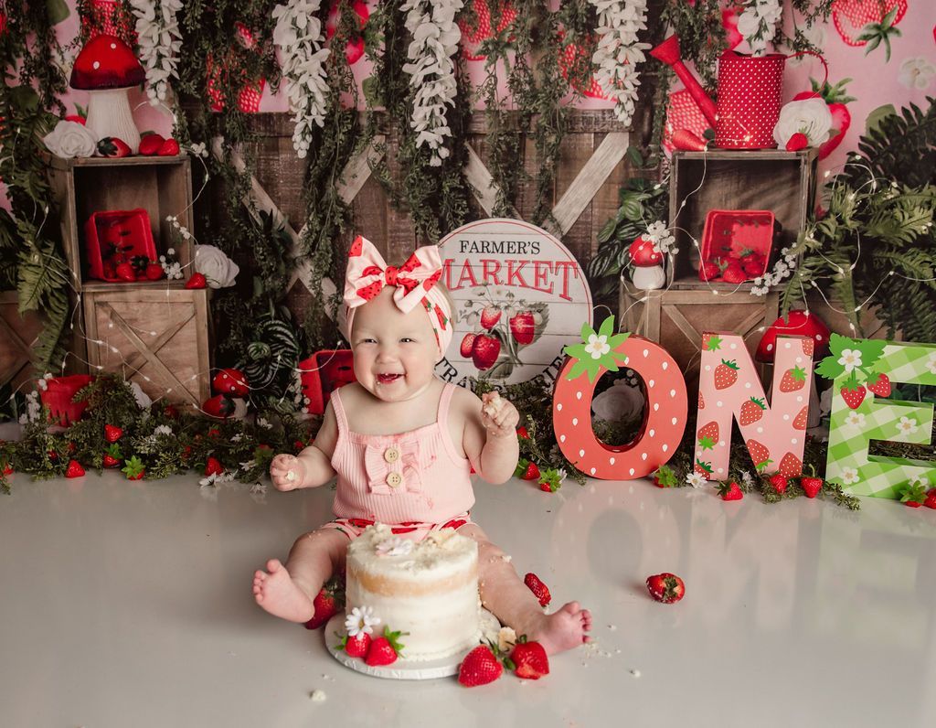 Happy baby in strawberry-themed setting, sitting near cake and the word 