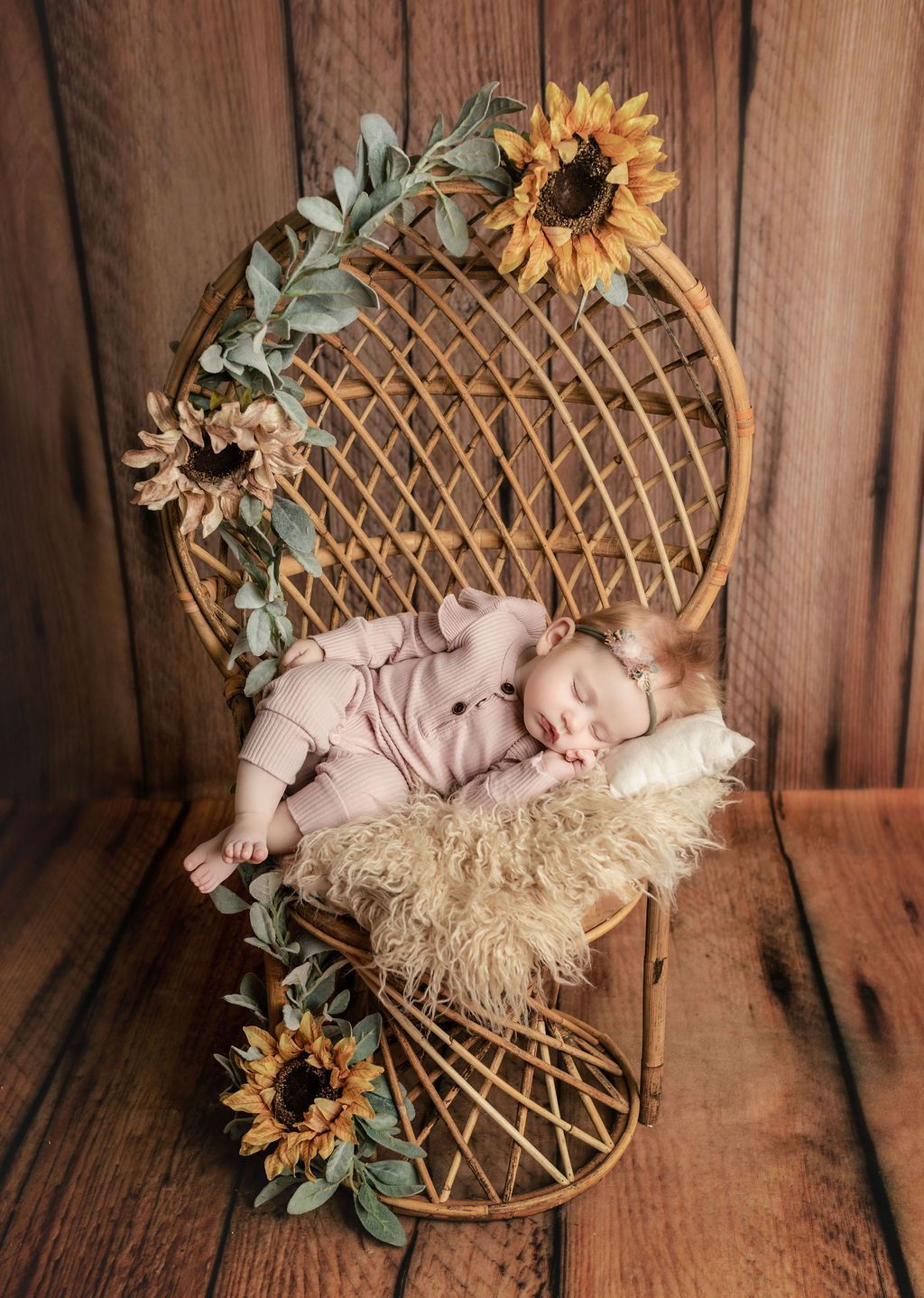 Newborn asleep in wicker peacock chair with sunflowers and greenery, rustic wood backdrop.