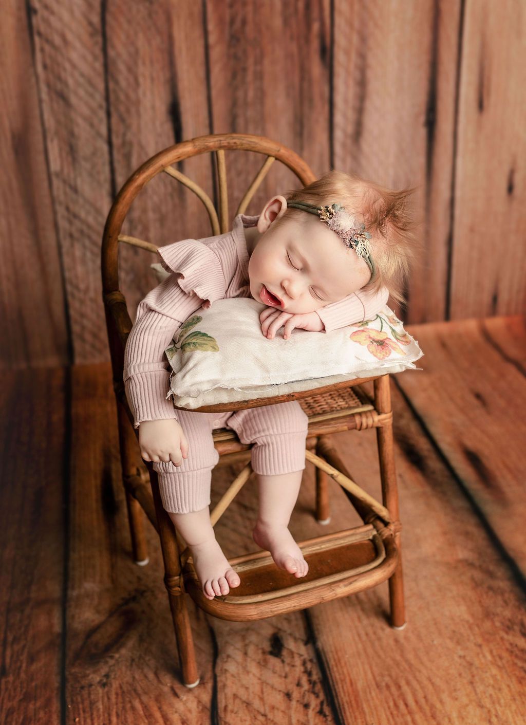 Baby sleeping in miniature wooden chair, resting head on pillow. Pink outfit, wood background.