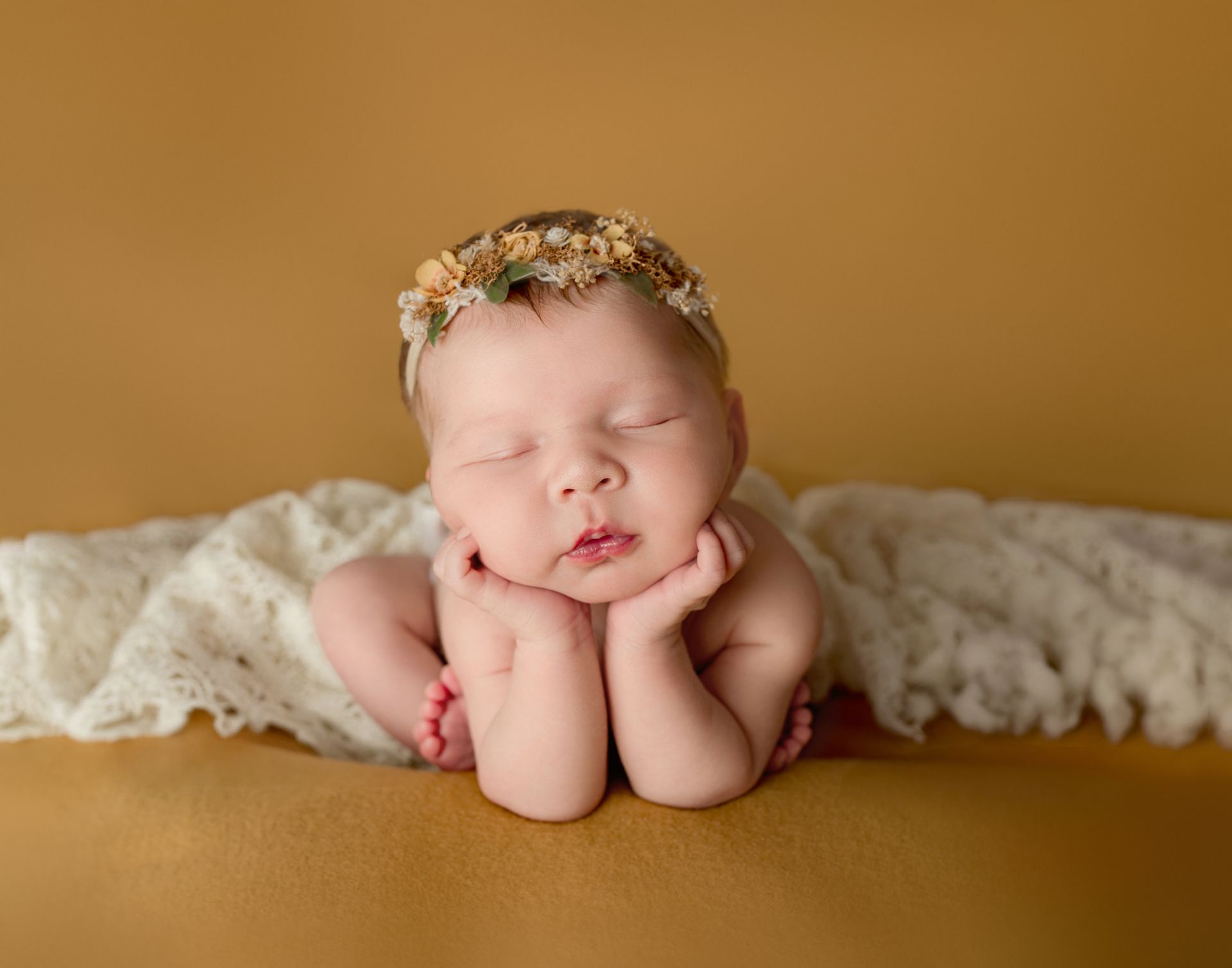 Newborn baby resting on hands, wearing flower crown, on a cream blanket, gold background.
