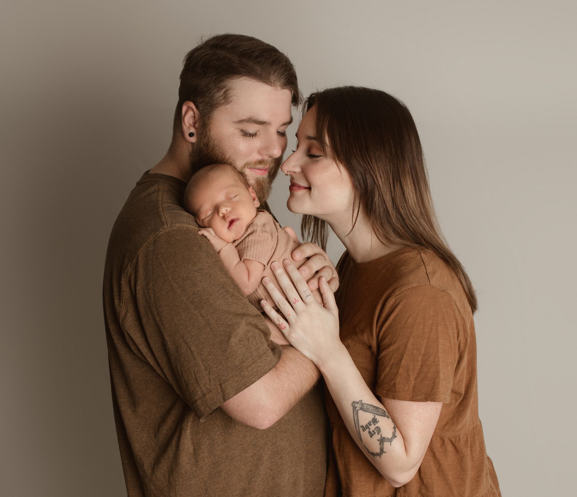 Parents holding newborn baby, smiling close together. Brown tones, plain backdrop.