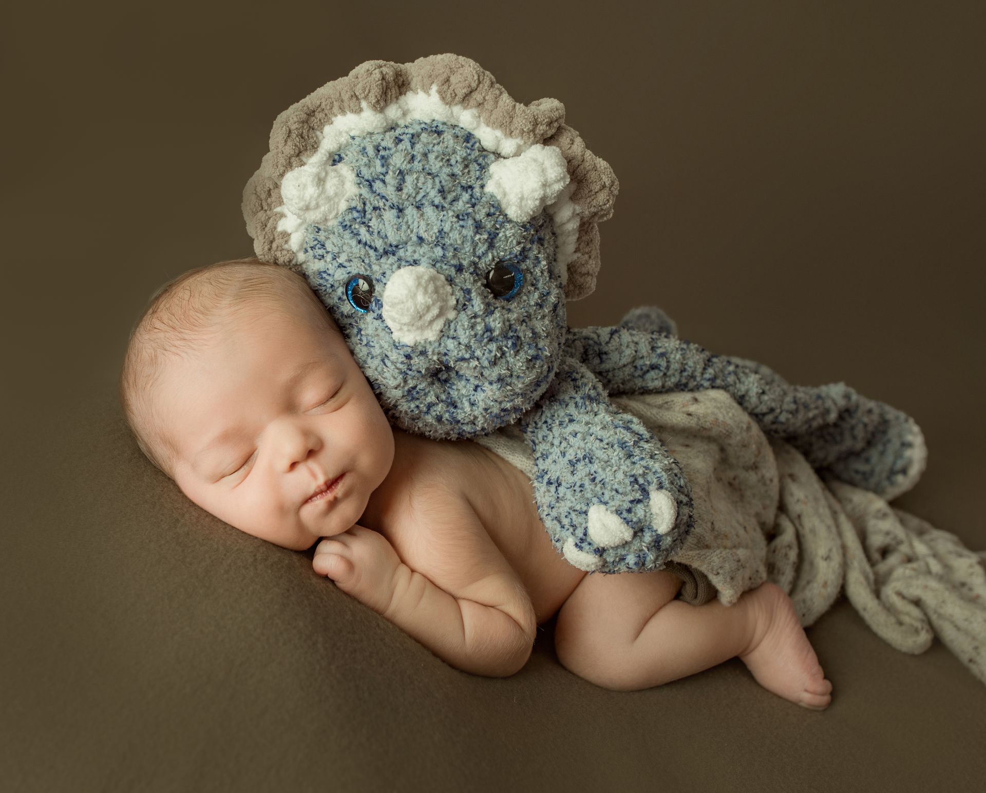 Newborn baby sleeping next to a blue and tan crocheted triceratops.