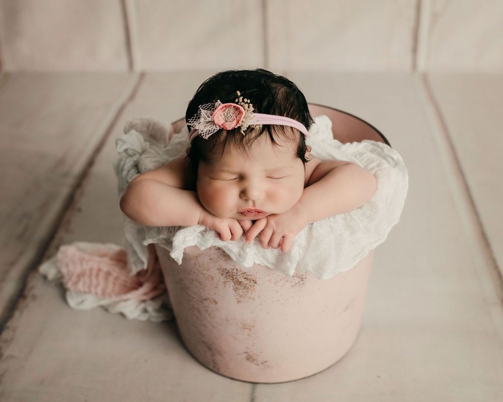 Newborn in a pink bucket, wearing a floral headband, sleeping peacefully on a white wrap.