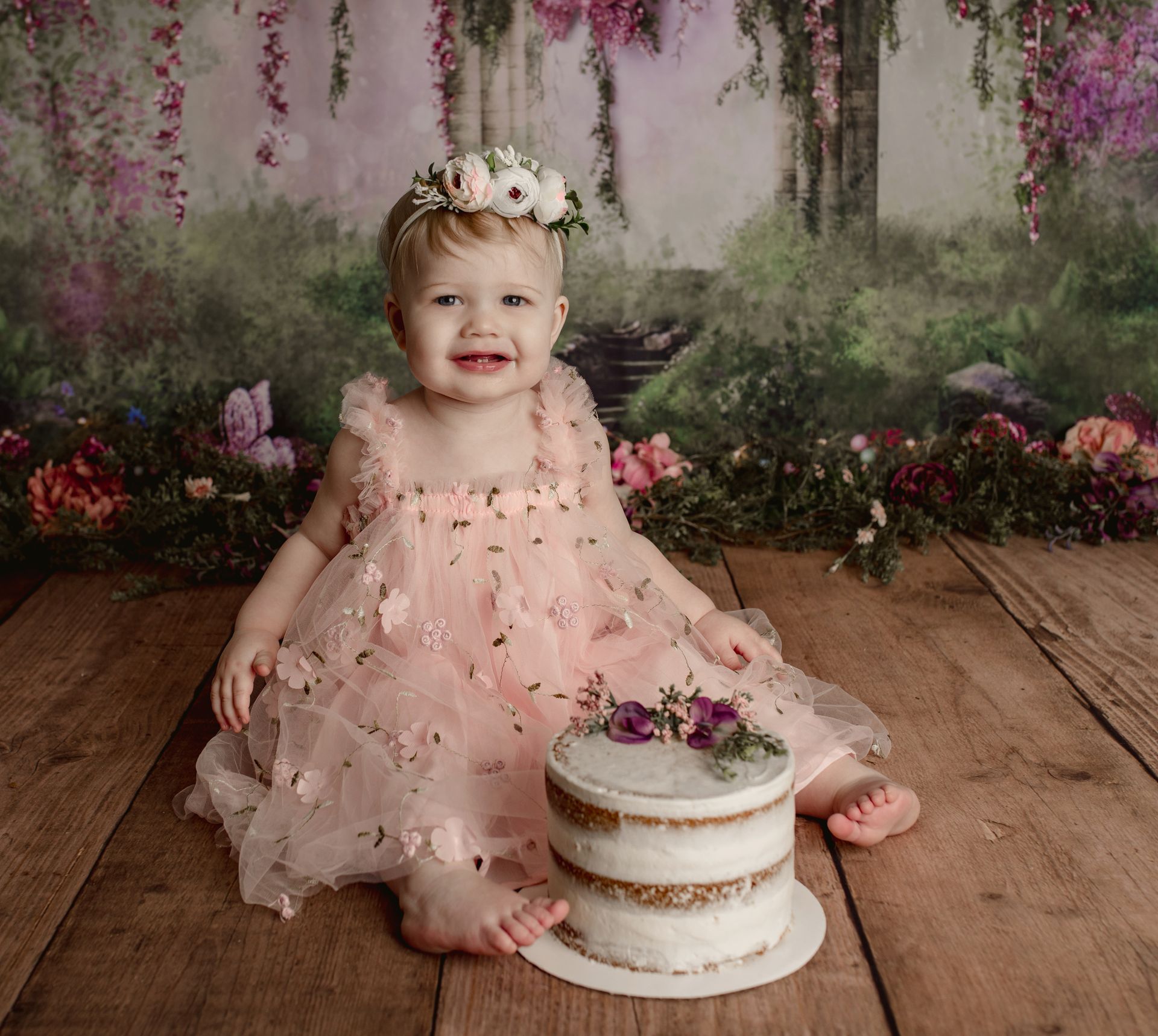 Girl in pink dress with floral accents smiles at a cake, wood floor, floral backdrop.