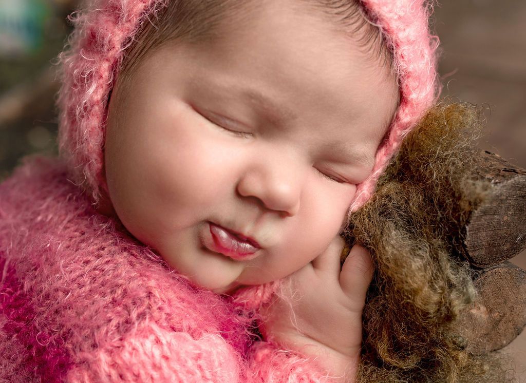 Sleeping baby wearing a pink knit hat and sweater, head resting on a fuzzy brown texture.