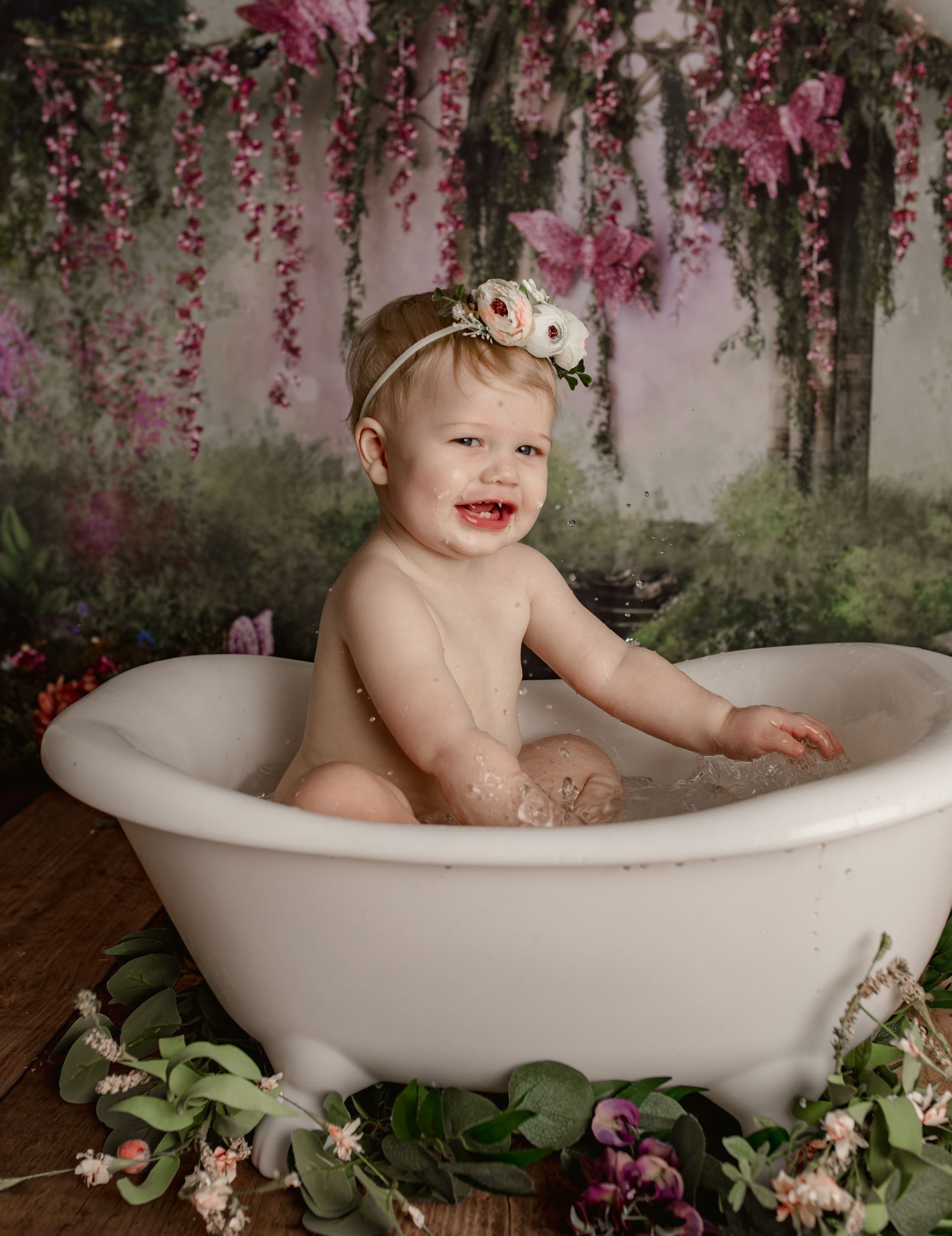 Baby in a clawfoot tub smiles, wearing a floral headband. Fairy garden backdrop with pink butterflies.
