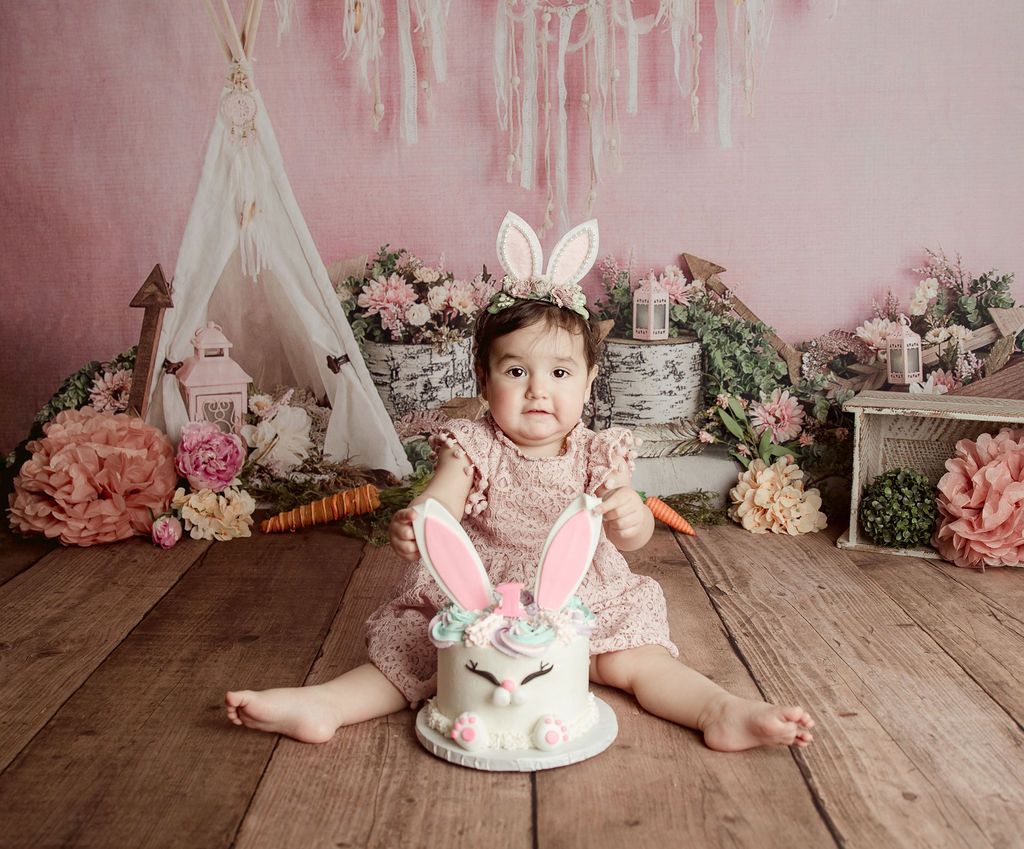 Baby wearing bunny ears, seated in front of a bunny-themed cake, pink background, rustic wood floor.