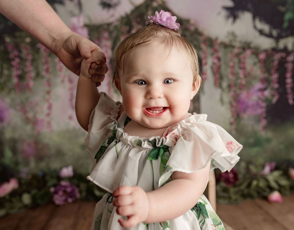 Smiling baby holds a hand, wearing floral dress, with a flower in her hair. Background includes flowers.
