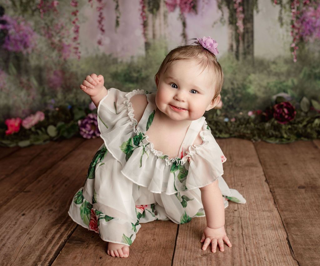 Smiling baby crawling on wooden floor, wearing a white floral outfit, with a floral backdrop.