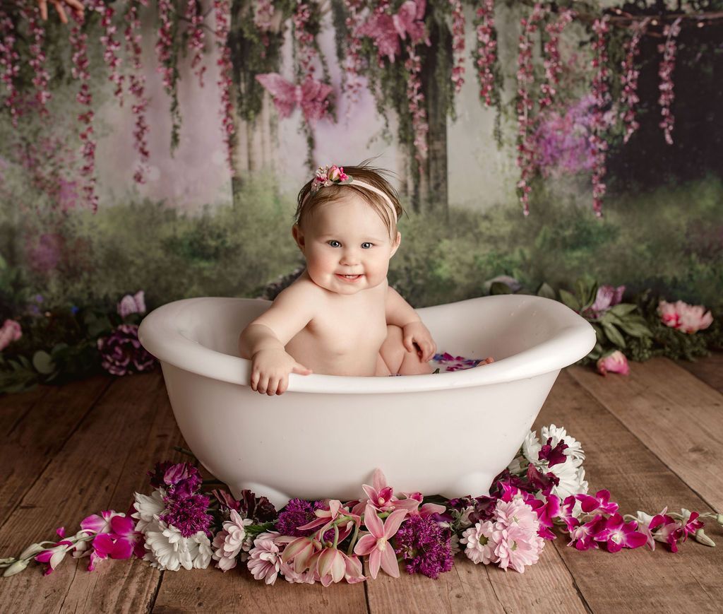 Baby in a white bathtub, surrounded by flowers on a wood floor, with a floral backdrop.