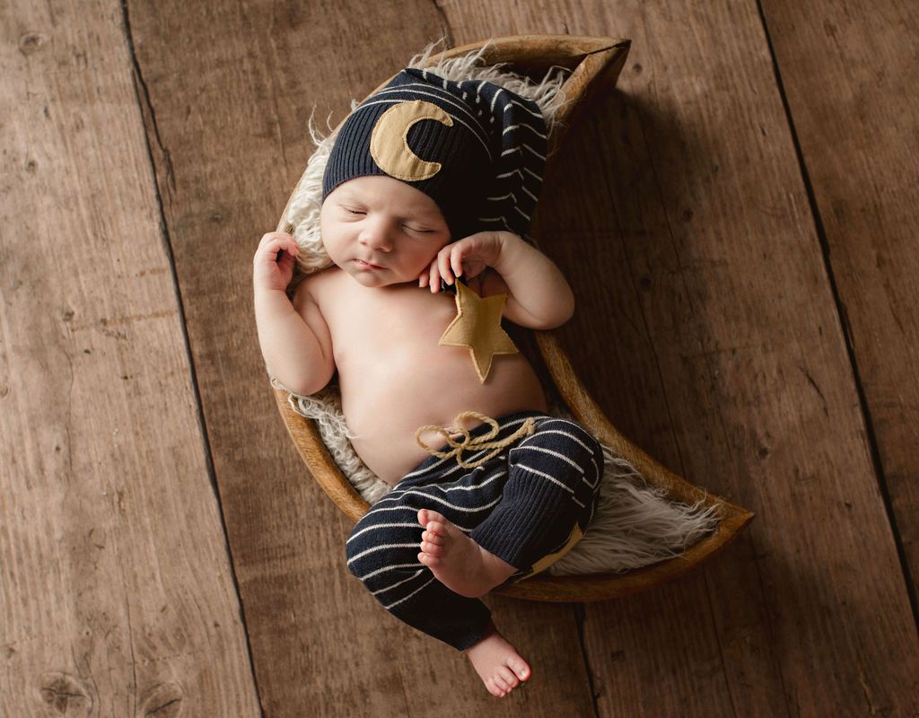 Newborn baby in crescent moon-shaped prop, wearing a moon and star hat and striped pants. Brown wooden floor.