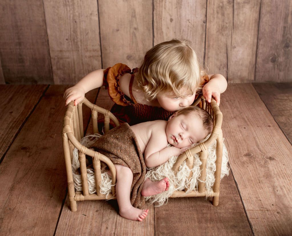 Toddler kisses sleeping infant in a small wooden bed; rustic wood backdrop.